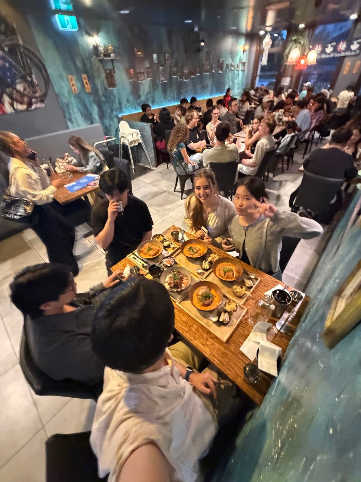 A selfie over a long table of friends sharing dinner in a busy restaurant