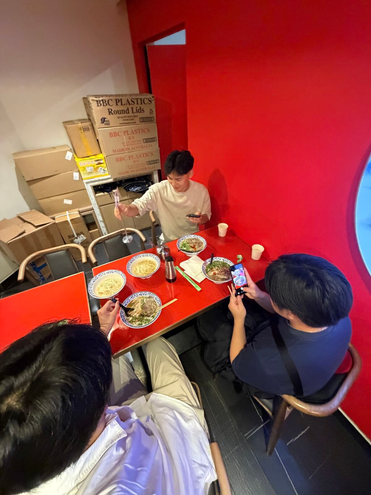 Eric and two friends with bowls of noodle soup at a red table, boxes stacked behind them