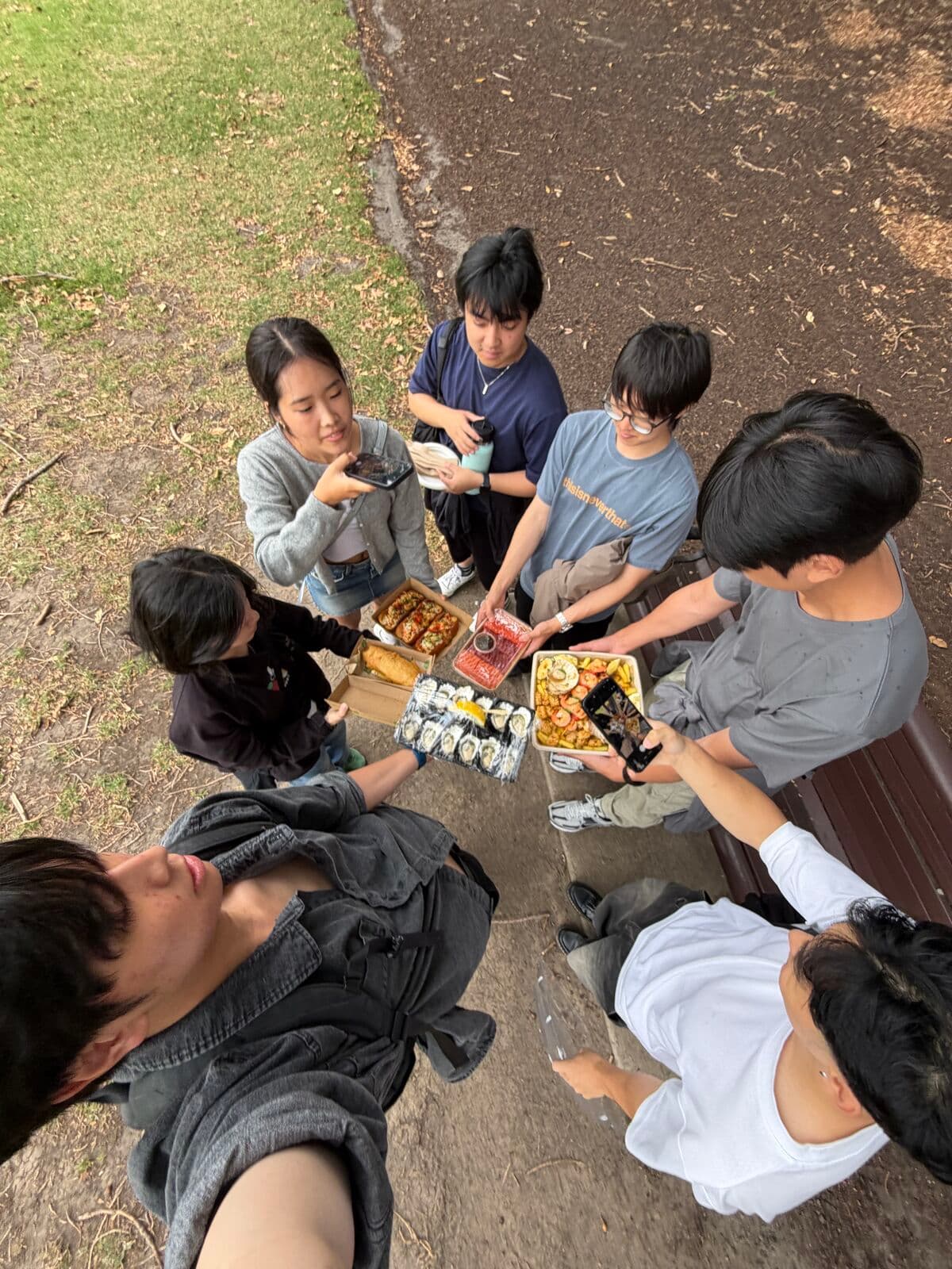 Friends standing in a circle by a park bench, holding out trays of sushi and snacks