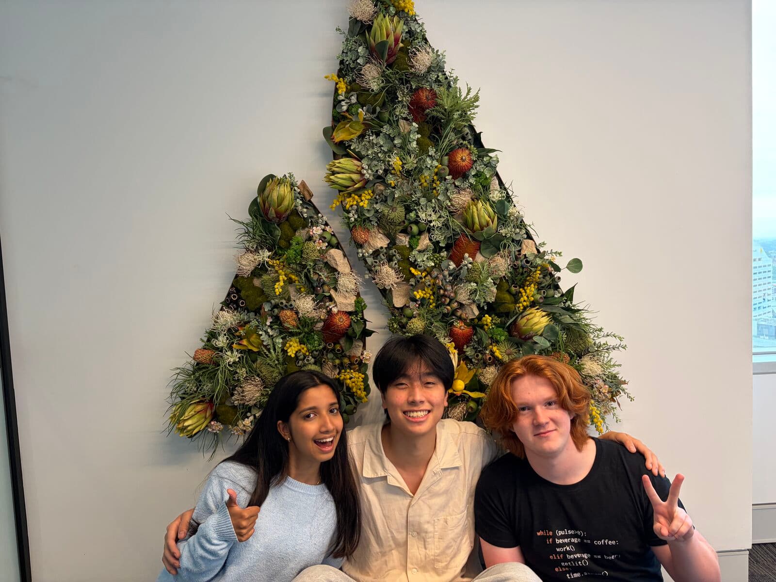 Eric and two friends sitting under a large triangle of native flowers on a white wall