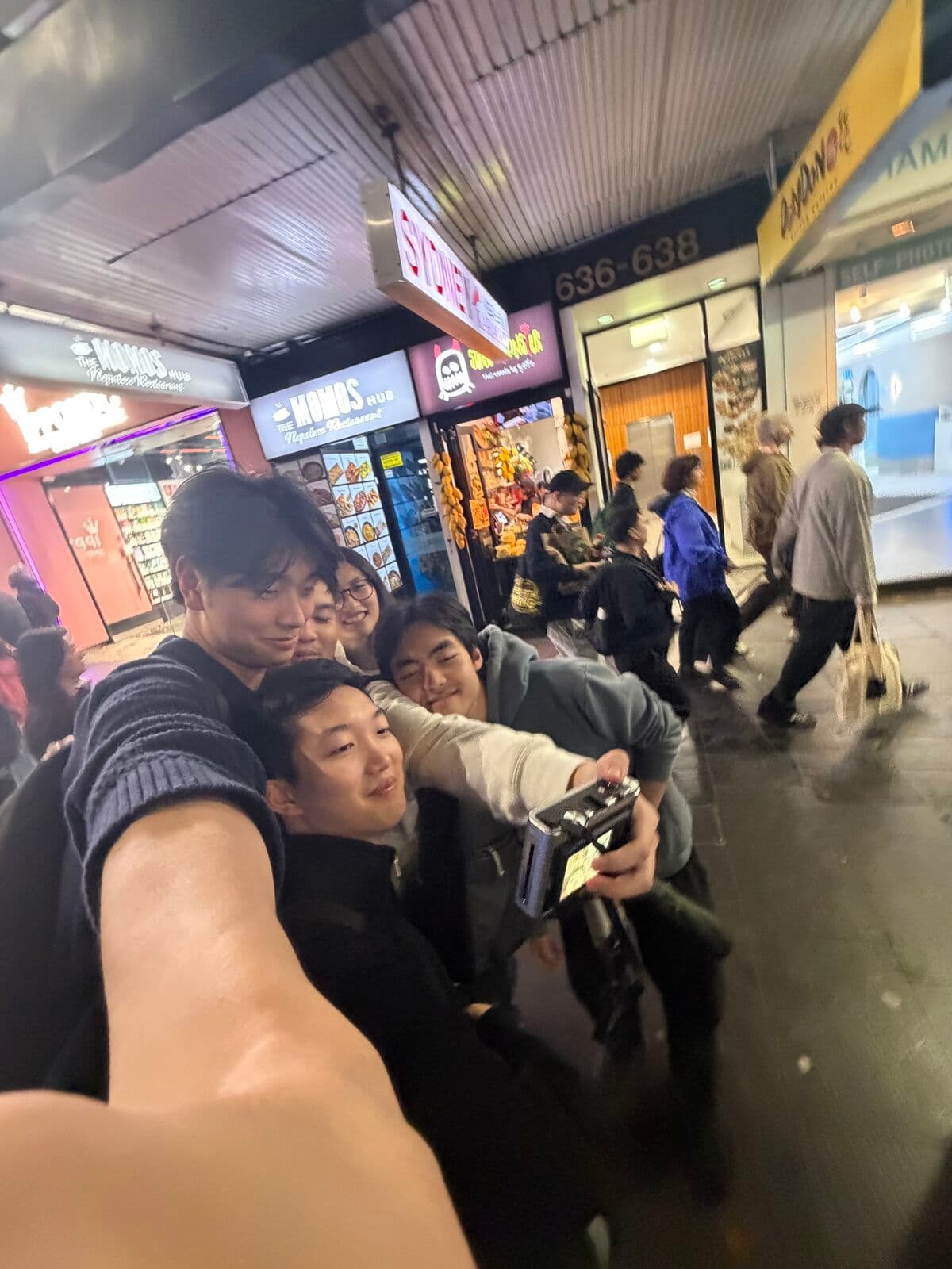 A selfie of five friends squeezed together outside shops on a busy street at night, one holding a camera