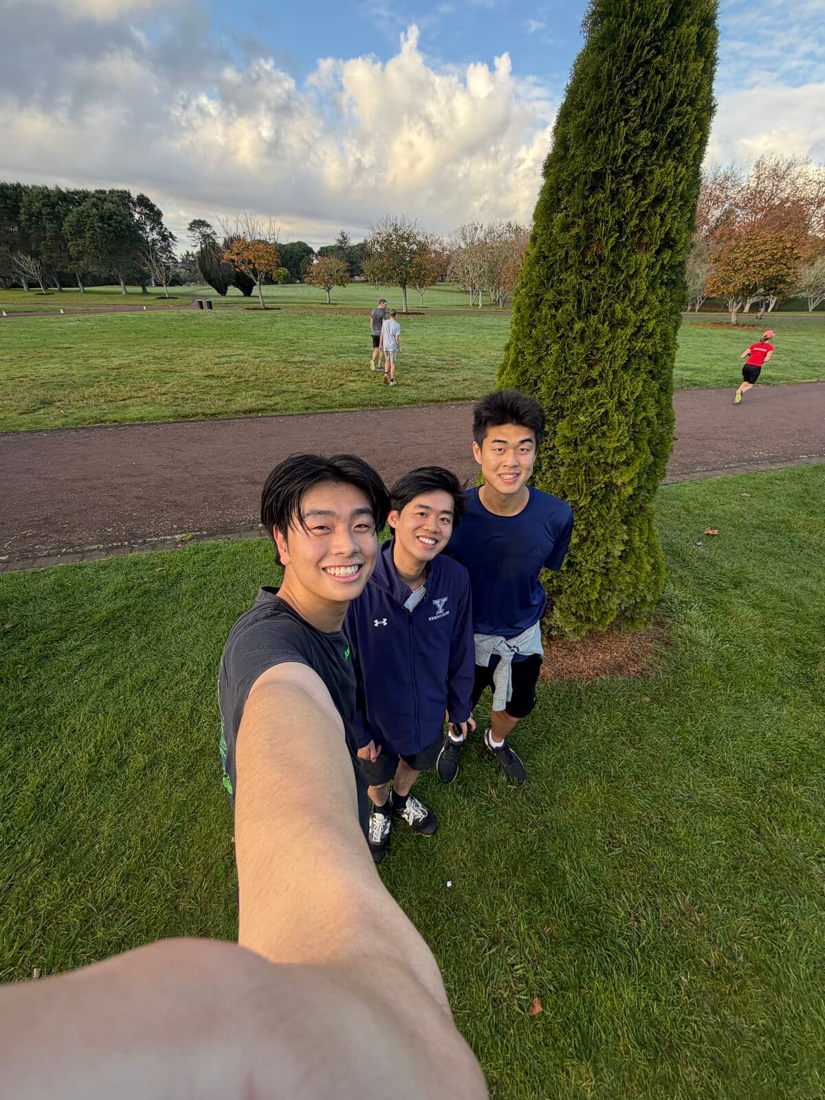 A selfie of three friends in running clothes on the grass of a park, beside a tall cypress tree