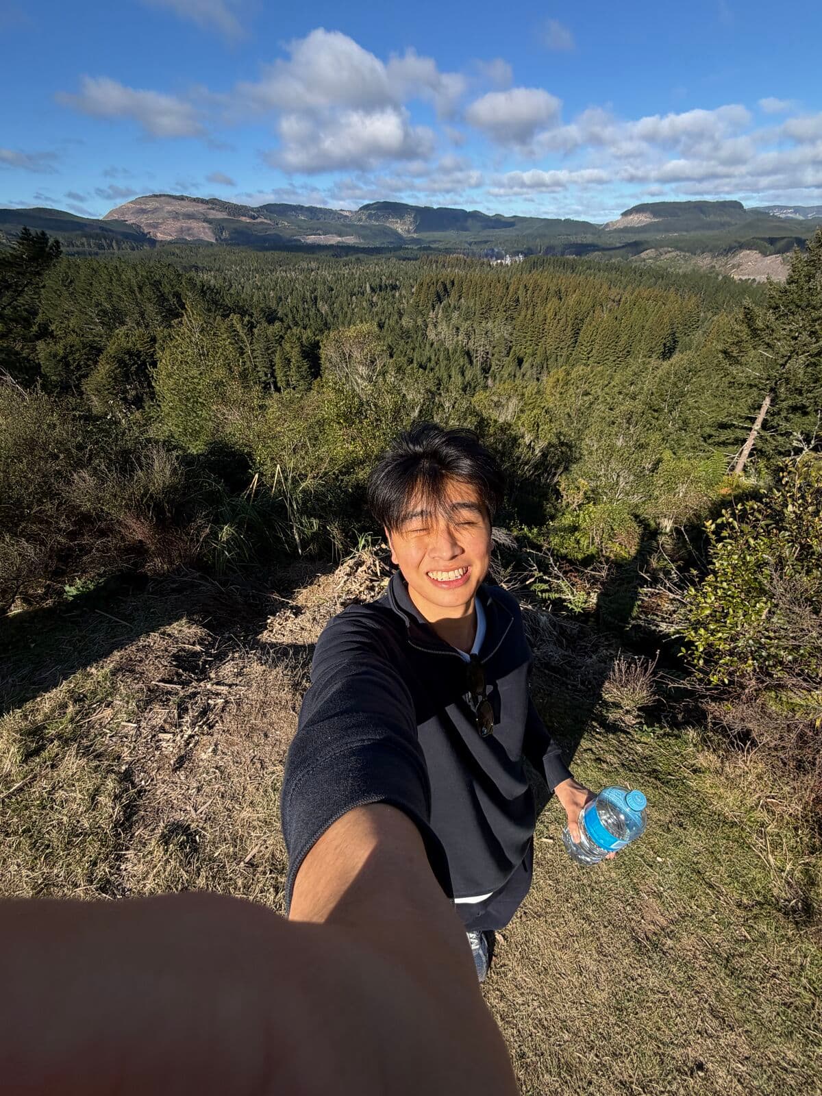 Eric taking a selfie on a hilltop above a forested valley, holding a water bottle