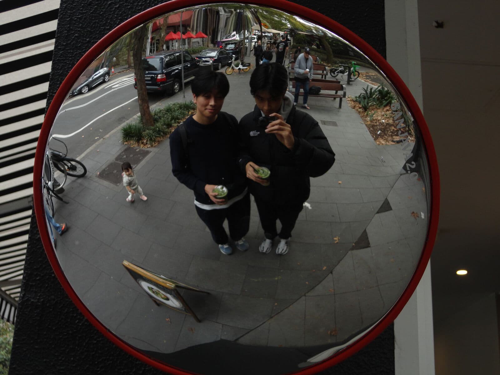 Eric and a friend holding iced drinks, reflected in a round mirror on a city street